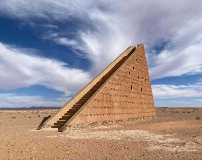 A large, triangular stone structure with a staircase leading up to its peak, set in a barren landscape under a partly cloudy sky. 3