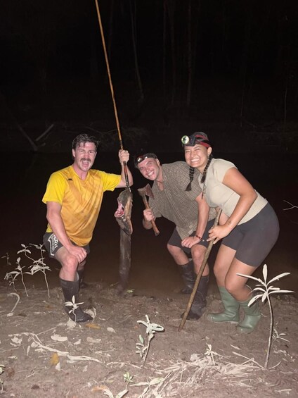 Three people holding a fish in a muddy outdoor setting at night. 3