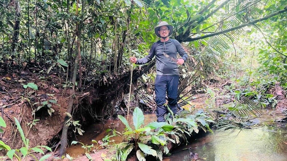 A person standing on a muddy bank in a forested area with a stream. 5