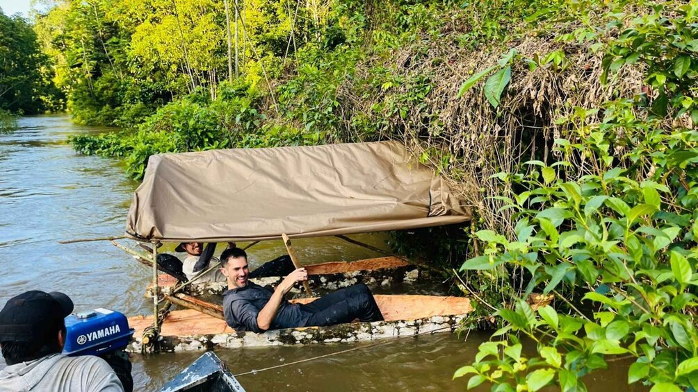 A makeshift shelter on a riverbank, a person lying under it, and another person in a boat nearby. 1