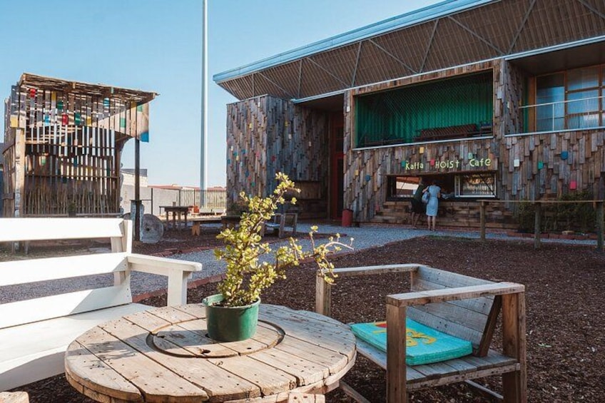 En utomhusplats med sittmöbler i trä, bestående av ett bord och stolar, en krukväxt och en byggnad med en unik träfasad och en skylt med texten ”Kaffee Hoist Cafe”. 3