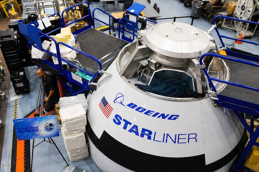 Un vehículo aeroespacial grande con la leyenda 'Boeing Starliner', rodeado de barandillas de seguridad azules y varios equipos. 4