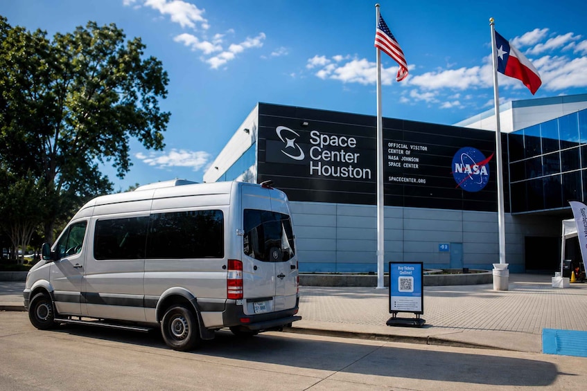 Una camioneta blanca estacionada frente al edificio del Centro Espacial de Houston, con banderas y el logotipo de la NASA. 2