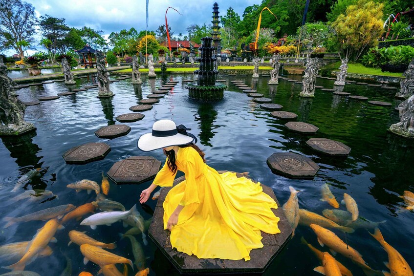 A woman in a yellow dress standing on a floating platform surrounded by koi fish in a pond with stone steps and a pagoda in the background. 4