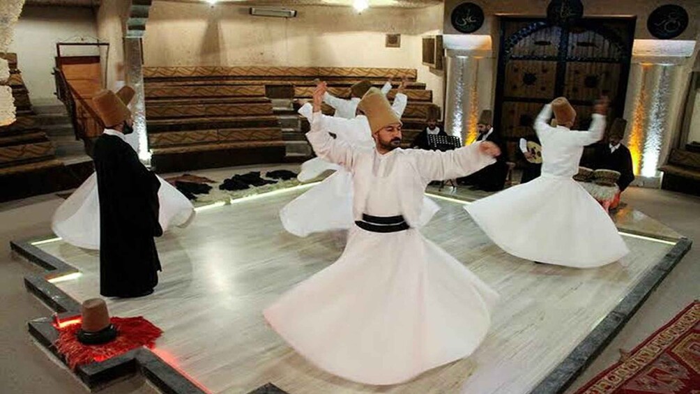 A person in white traditional attire performing a dance in a room with a patterned carpet and a wooden bench. 4