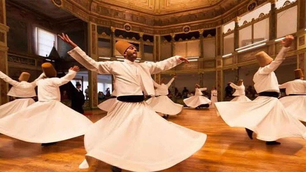 A group of people in white traditional attire performing a dance in a grand hall with ornate architecture. 2