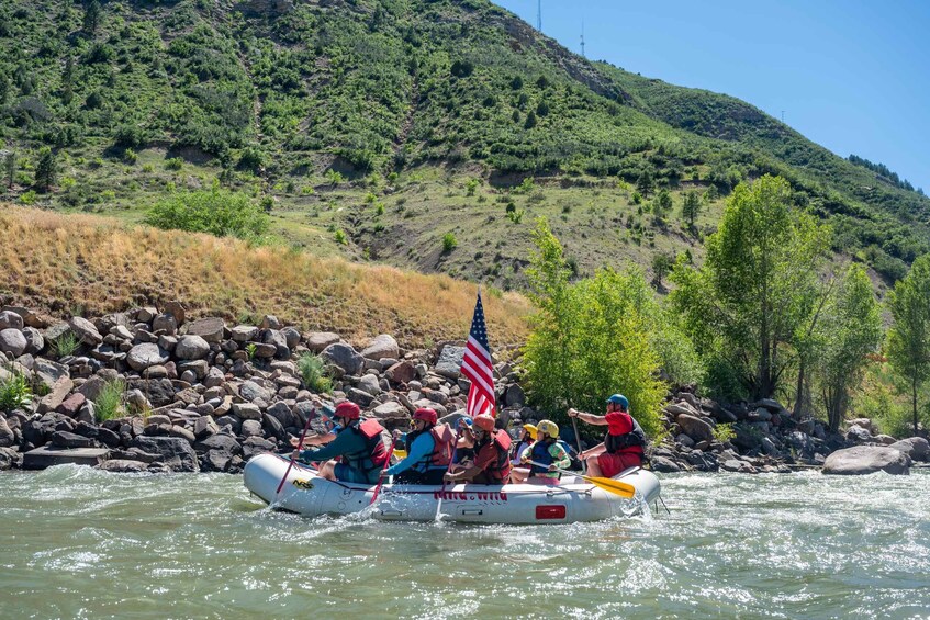 A group of people rafting down a river, wearing life jackets and helmets, with an American flag on the raft. 1