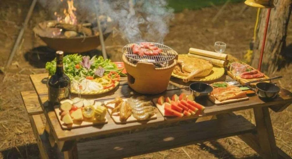 A wooden table with various foods, a grill, and a bottle of drink outdoors at night. 5