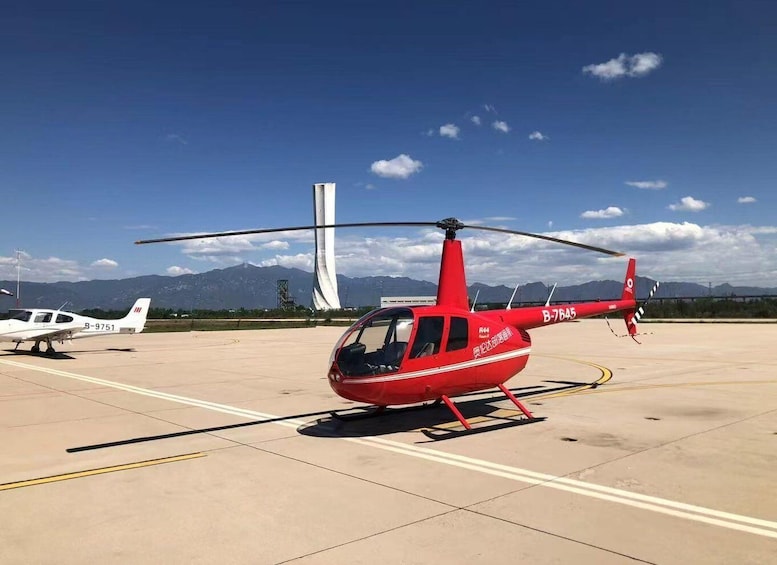 A red helicopter with registration B-7645 on a tarmac, a white airplane in the background, and mountains in the distance. 2