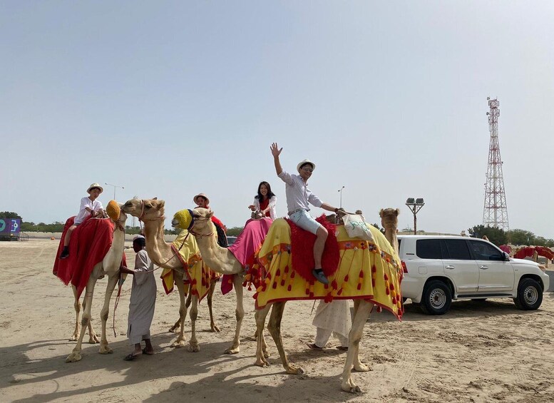 A group of people riding decorated camels on a sandy beach with a white car and a tall communication tower in the background. 1