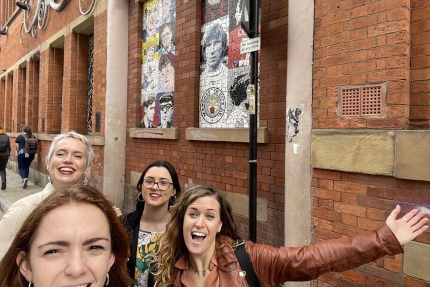Eine Gruppe von Personen macht ein Selfie vor einem Gebäude mit einem Wandgemälde und einer Backsteinmauer. 1