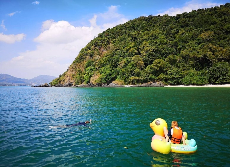 A person swimming and two people on inflatable rafts in a clear blue sea with a forested island in the background. 3