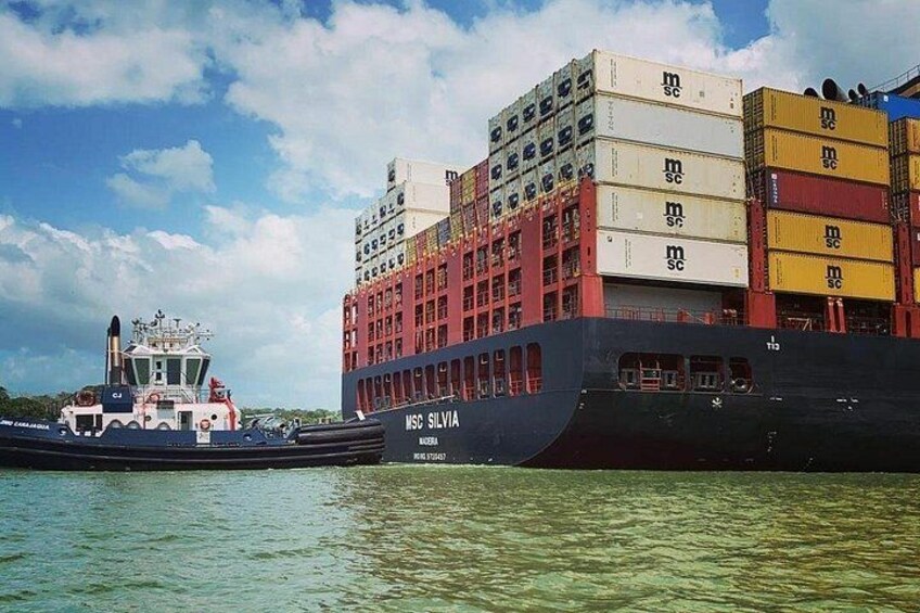 Ein großes Frachtschiff mit gestapelten Containern, ein Schlepper und ein klarer Himmel. 2