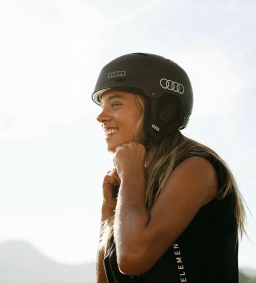 A person wearing a helmet with a logo, standing outdoors with mountains in the background. 3