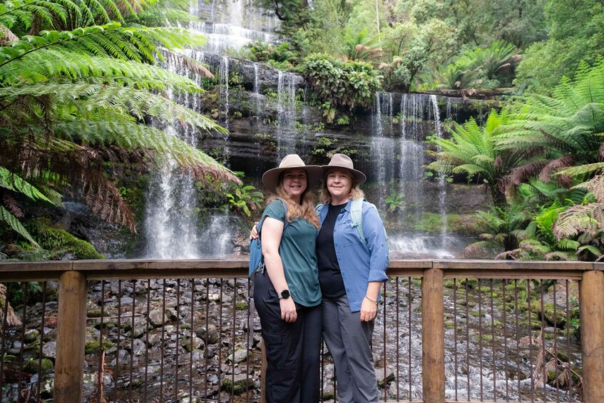 Two people standing in front of a waterfall with lush greenery and ferns. 1