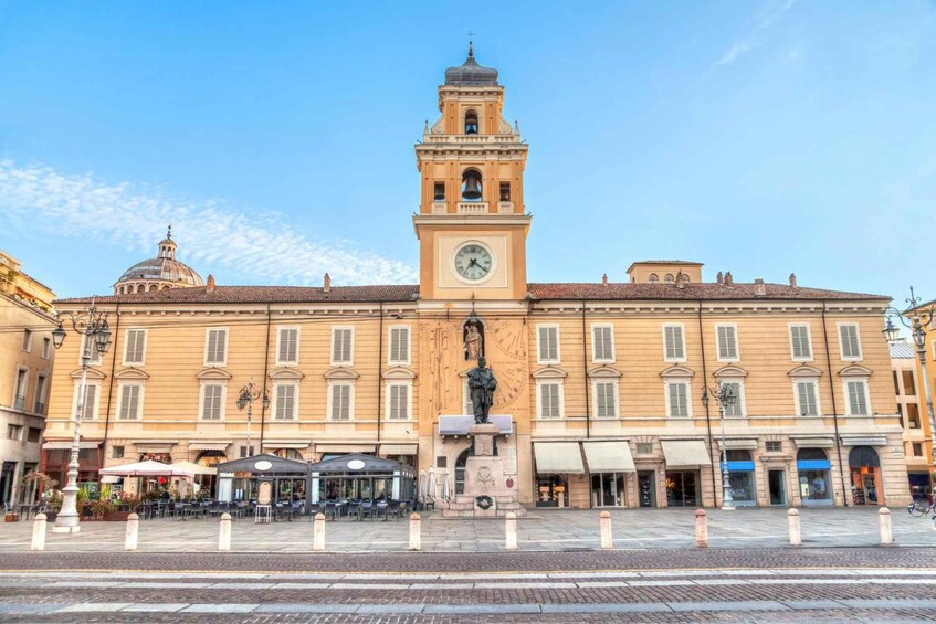 A historic building with a clock tower, a statue in front, and a plaza with bollards. 5