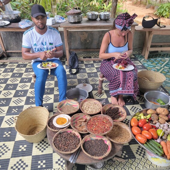 Two people sitting on a patterned mat with food and spices in front of them. 3
