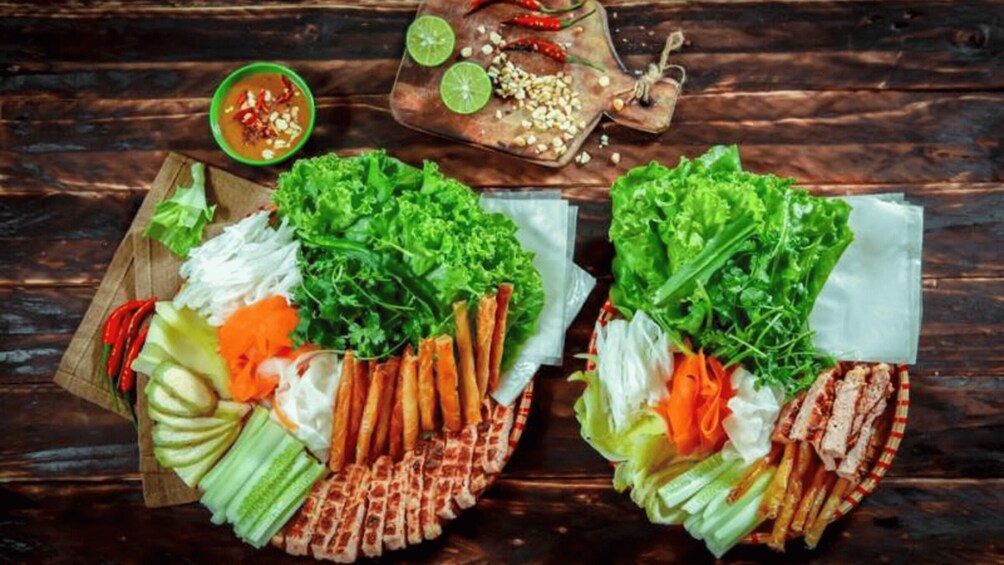 A wooden table with two plates of fresh vegetables and herbs, a bowl of dipping sauce, and a small dish with chili peppers and lime. 4