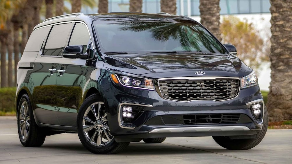 A dark-colored Kia minivan parked outdoors with palm trees in the background. 1