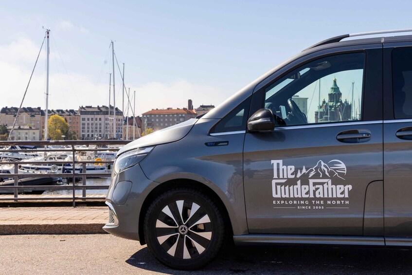 A silver car with 'The GuideFather' logo parked by a marina with boats and buildings in the background. 1