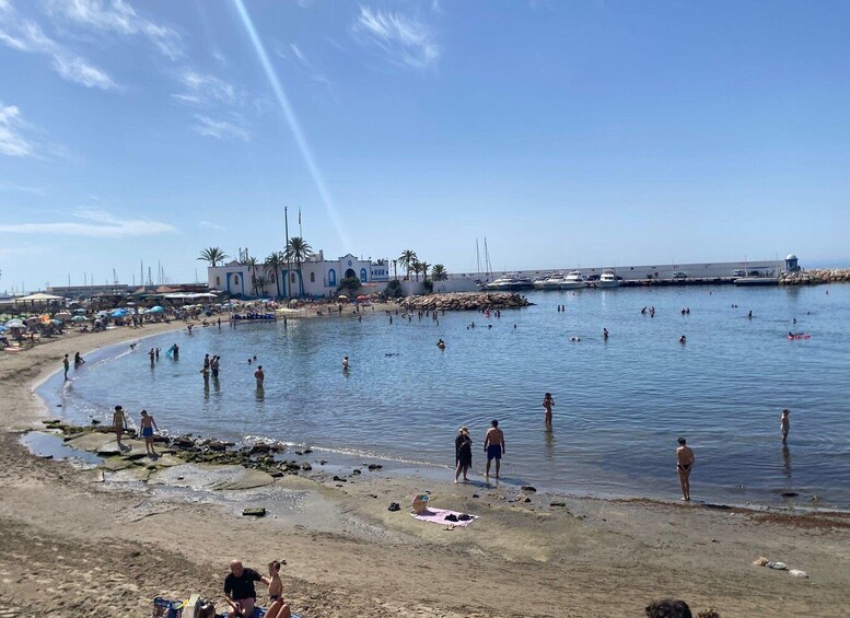 A beach with people swimming and sunbathing, a marina with boats, and a building with a blue and white facade. 4