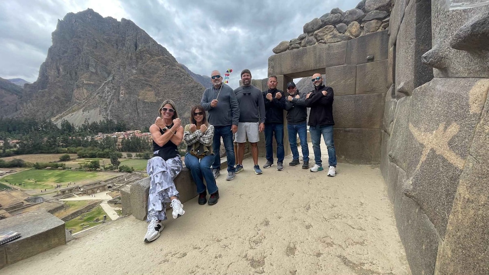 A group of people posing in front of a stone structure with a mountainous landscape in the background. 2