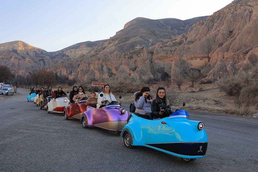 A group of people riding colorful go-karts on a road with a mountainous backdrop. 1