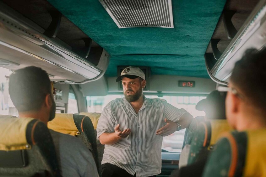 A man gesturing in a bus interior with a digital clock displaying 6:03. 2