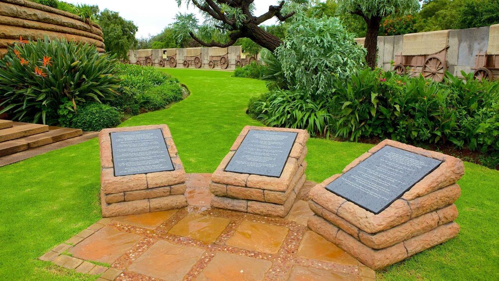 A garden with stone plaques on a stone pathway, surrounded by lush greenery and a large tree. 1