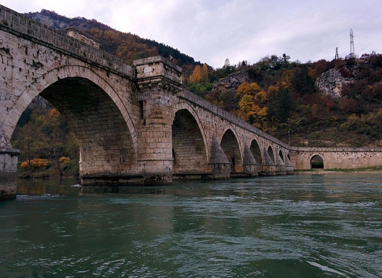 A stone bridge over a river with multiple arches, surrounded by autumn foliage and hills. 2
