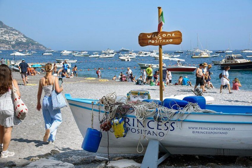 Een strand met mensen die wandelen, een bord waarop staat 'Welcome to Positano' en boten in het water. 5