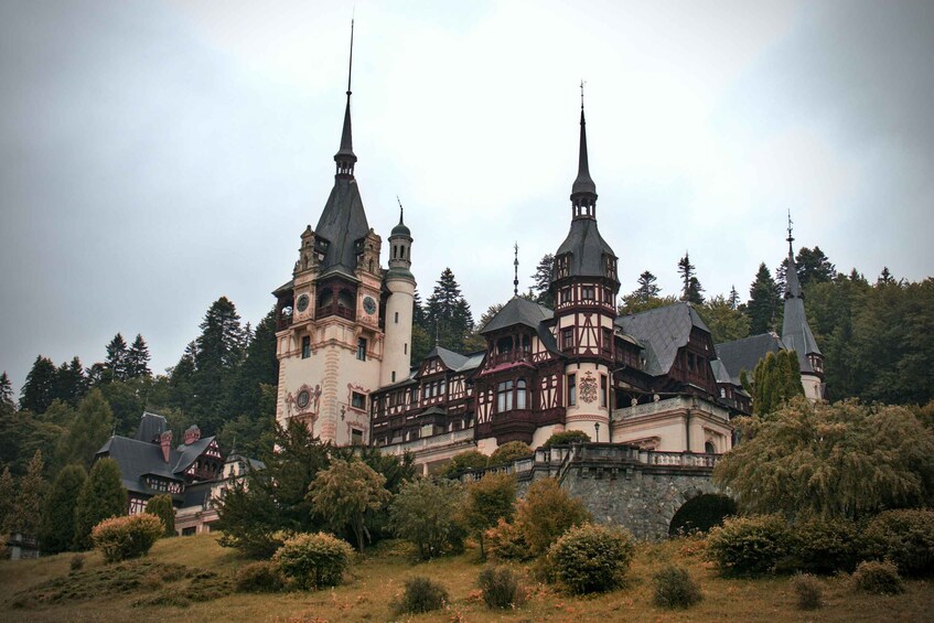 A historic castle with multiple towers and a large clock tower, surrounded by lush greenery and trees. 1