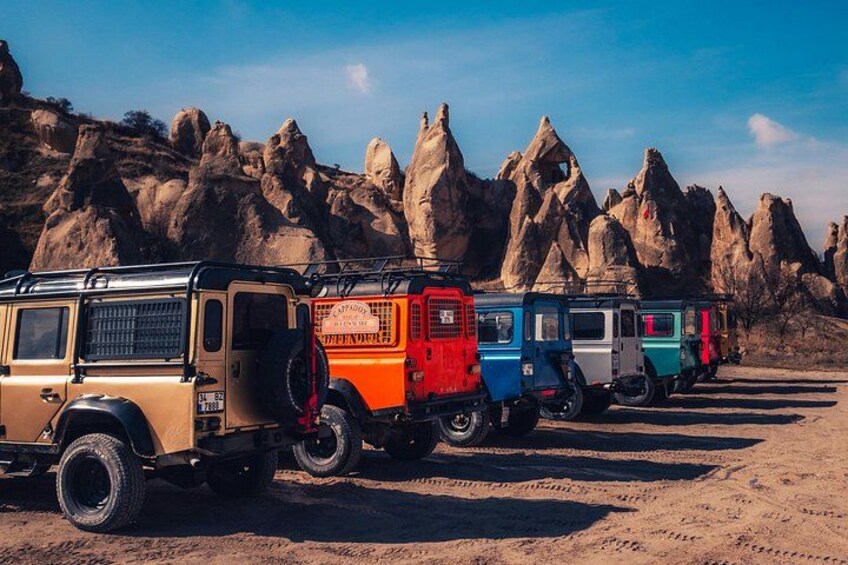 A row of colorful vehicles parked on a sandy surface with rocky formations in the background. 5