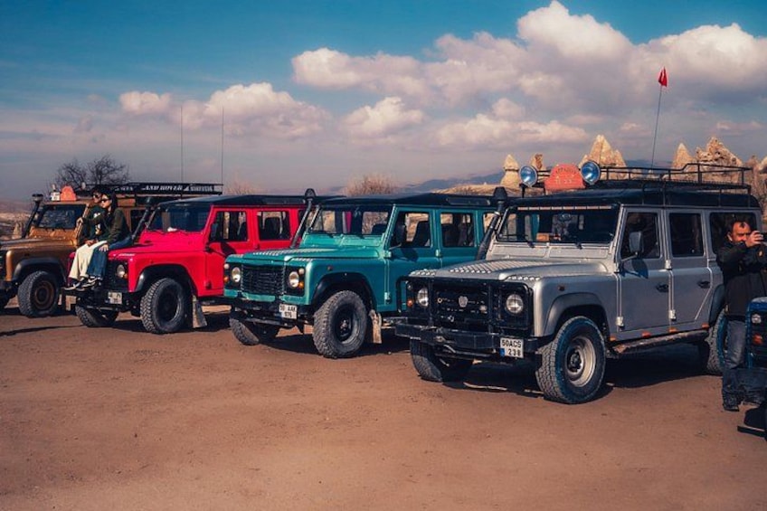 A row of off-road vehicles parked on a dirt surface with a mountainous landscape in the background. 1