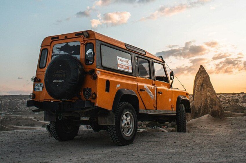 An orange off-road vehicle parked on a gravel surface with a rocky outcrop in the background. 2