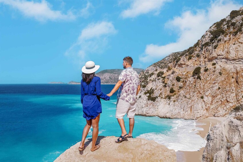 A couple standing on a rock by a turquoise sea with a rocky cliff in the background. 1