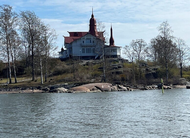 A large house with a red roof and a tall spire, surrounded by trees and a rocky shoreline. 3