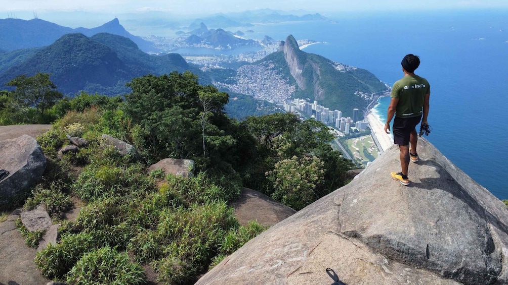 A person standing on a rocky outcrop overlooking a city and coastline. 1
