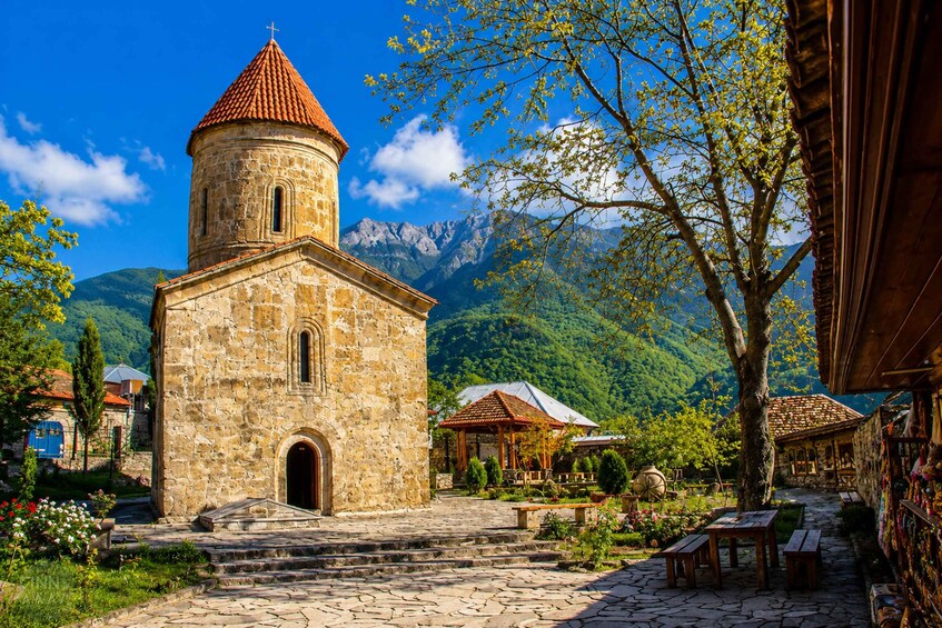 A stone church with a bell tower, surrounded by a cobblestone courtyard, trees, and mountains in the background. 1