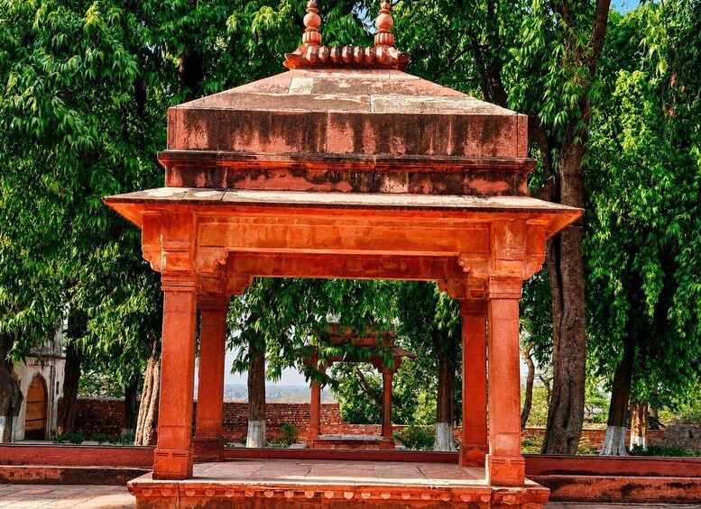 A red stone pavilion with intricate carvings, surrounded by lush green trees. 2