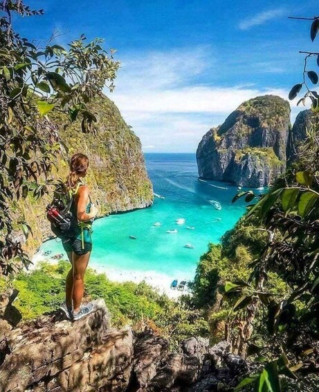A person standing on a cliff overlooking turquoise waters with boats, surrounded by lush greenery and rocky cliffs. 1