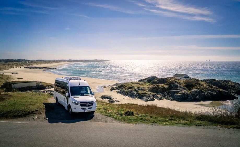 A white van parked on a coastal road with a beach and rocky outcrops in the background. 4
