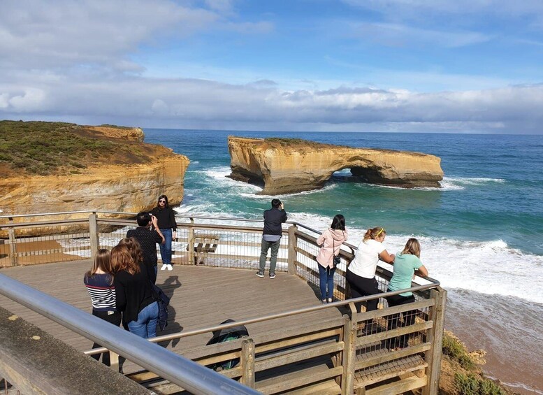 A coastal scene with a wooden boardwalk, people observing the ocean, and a prominent rock formation with a natural arch. 4