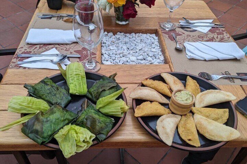 A table with tamales and empanadas, a bowl of sauce, and a vase with flowers. 2