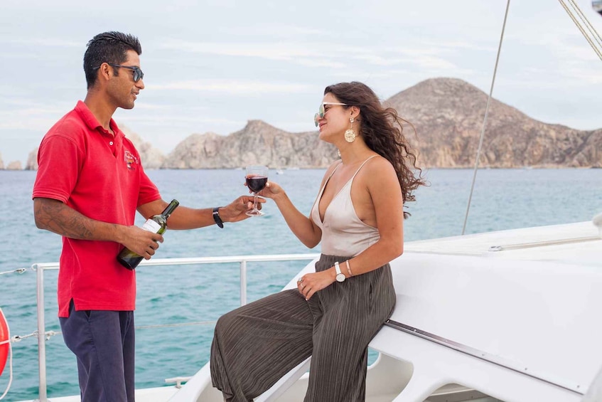 A man and a woman on a boat, holding a bottle and a glass of red wine, with a mountainous coastline in the background. 4