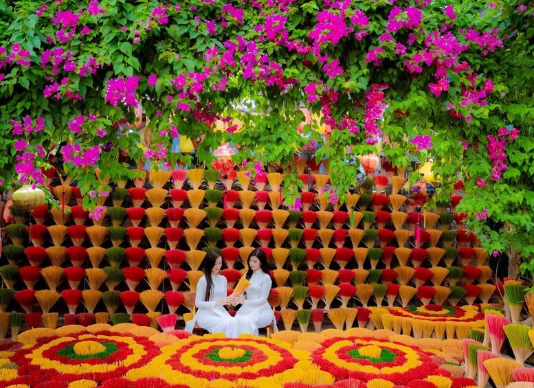 A woman in a white dress sitting on a colorful floral carpet, surrounded by vibrant flowers and greenery. 2