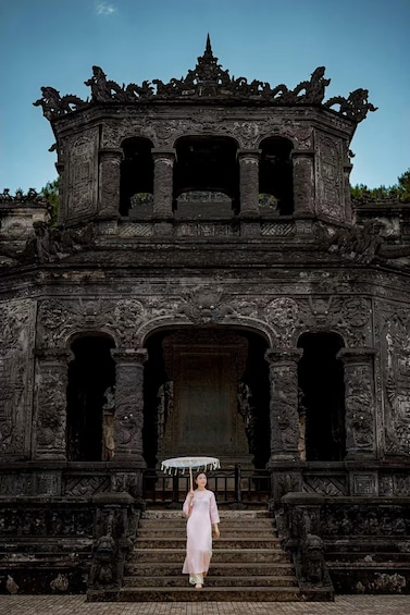 A woman in a pink dress walking up a set of stone steps towards an ornate temple entrance with intricate carvings and a traditional umbrella. 3