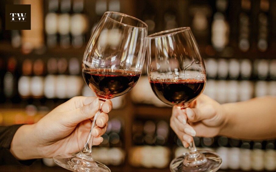 Two hands holding wine glasses filled with red wine, with a blurred background of bottles. 1