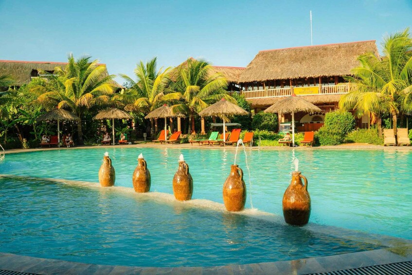 A swimming pool with water jets, palm trees, and thatched-roof buildings. 1