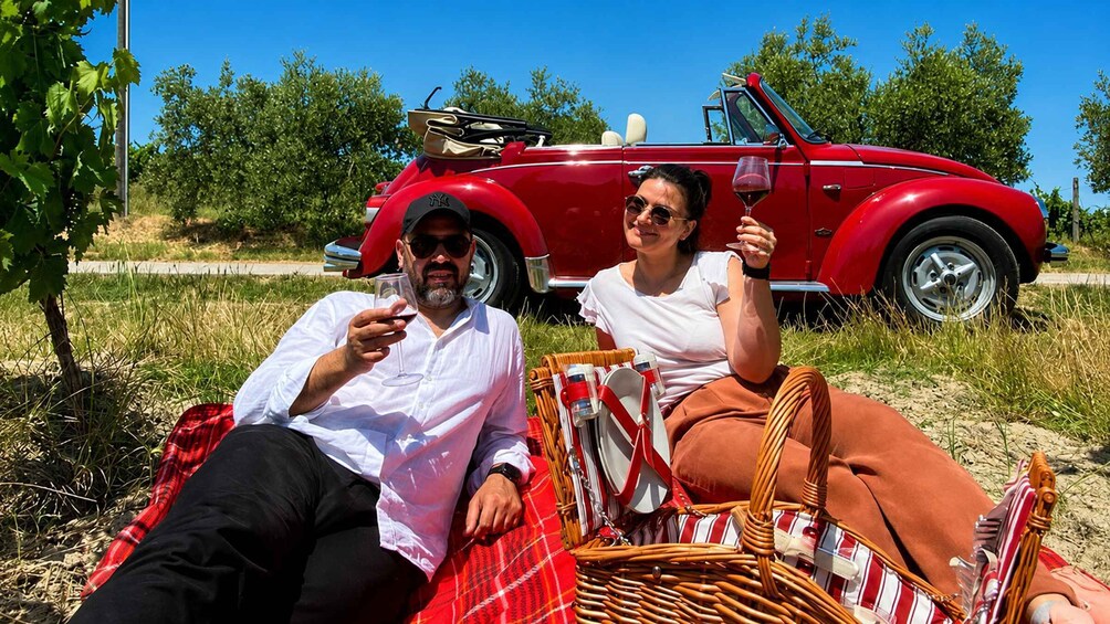 Two people relaxing on a red and white striped blanket in a grassy field with a vintage red convertible car in the background. 3